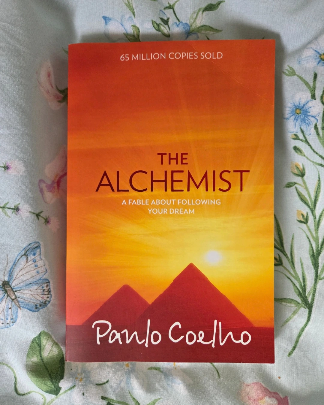 An image of the front cover of The Alchemist by Paulo Coelho. On orange shy with orange pyramids and black text